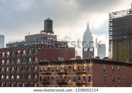 Older brownstone building are in the forefront with the New York City skyline in the background. A wooden water tank sits on top of one of the brick buildings. A building under construction is behind.