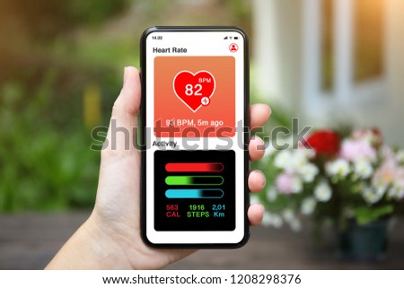 female hand holding phone with app heart and activity screen on background of house with flower garden