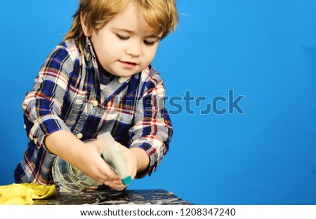Boy in checkered shirt on blue background. Cleaning activities concept. Child with busy face holds spray above table. Kid near wooden table with rag and foam on.
