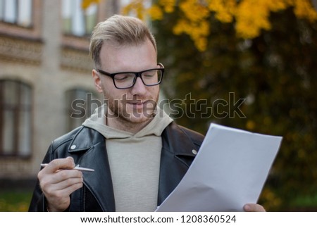 Portrait of a smiling student in casual clothes holding a pen with papers and looking at them, a young pen in black glasses offers to sign documents