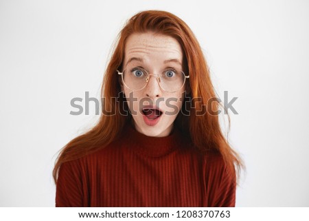 Picture of nerdy young woman with long ginger hair opening mouth in shock, seeing something surprising. Attractive shocked bug eyed red haired girl in glasses posing in studio, overwhelmed by emotions