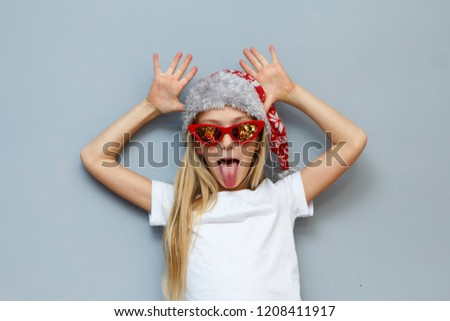 Christmas girl in Santa hat and sunglasses stylized sixties fools around with tongue sticking out on grey background
