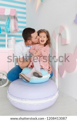 Young curly girl and lovely father posing sitting on funny big fake macaroon sweetness , candy shop style, pastel colors, positive emotions