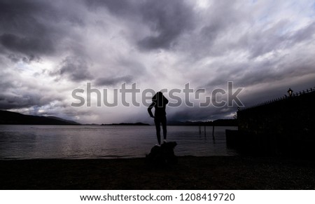 Girl on pedestal stares into the distance across bitter cold waters enjoying the picture perfect skyline as the tide laps against the shore. She is engulfed by a breathtaking cloudy sky overhead.   