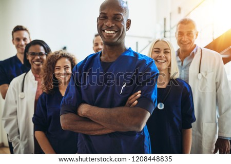 Young African male doctor smiling while standing in a hospital corridor with a diverse group of staff in the background Royalty-Free Stock Photo #1208448835