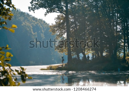 fishermen silhouettes on a bank of little natural lake in forest, foggy morning beautiful summer sunrise, backlight on tree leaves, still water surface, nature panorama background photo