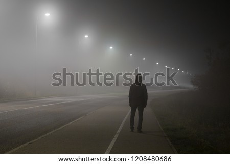 Young man alone slowly walking under white street lights in night. Dark time. Peaceful atmosphere in mist. Foggy air. Back view. Royalty-Free Stock Photo #1208480686