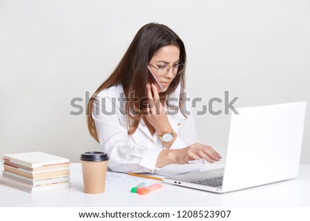 Skilled female student writes homework, works on laptop computer, uses application, keyboards and searches internet, has attentive look into screen, stay at table indoor, books and coffee near