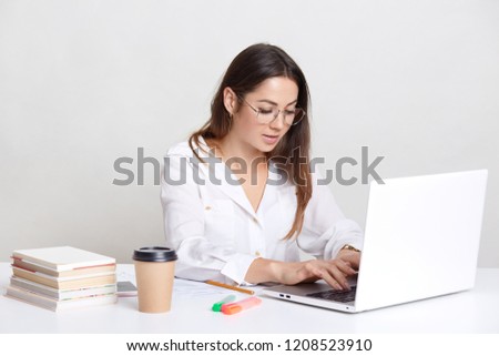 Busy freelancer recieves email, types feedback on laptop computer, wears round glasses, dressed in white shirt, surrounded with books, isolated over white background. People and career concept