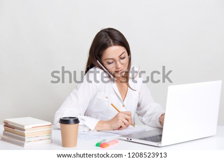 People, technology and business concept. Image of working woman has phone conversation, writes down some notes in notebook, searches information on laptop computer, drinks coffee, isolated on white