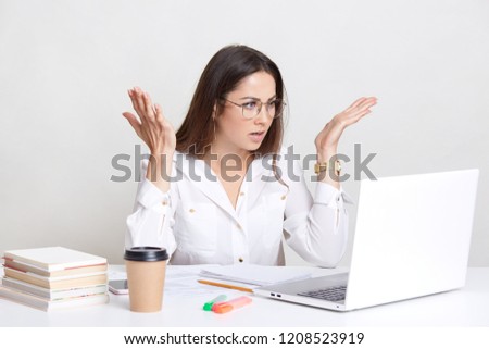 Puzzled emotive woman has deadline task, gesture with frustration, stares with bugged eyes, wears round spectacles for good vision, drinks coffee, isolated over white background. So what to do?