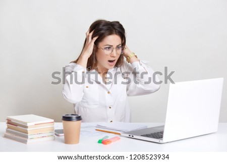 Puzzled young European female student in panic, stares with surprised expression into screen of laptop computer, has deadline task, drinks aromatic takeaway coffee, prepares for examination.