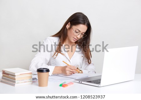 Female blogger wears white shirt, round spectacles, writes notes in notepad, works on laptop computer, drinks coffee, busy with developing webiste, uses wireless interent connection, isolated on white