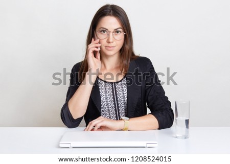 Female freelancer enjoys distant work at home, has telephone conversation, sits at white desk near laptop computer and glass of water, isolated over white background. People and technology concept