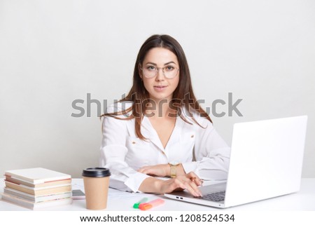 Succesful copywriter busy working with laptop computer, wears round glasses, drinks takeaway coffee, poses against white background, dressed in formal clothes. People, work and occupation concept