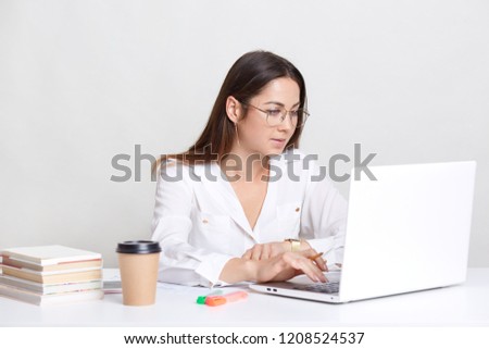 Young female administrative works on laptop computer, searches information in internet, dressed in white drinks hot beverage, uses books for writing diploma paper, isolated over white background