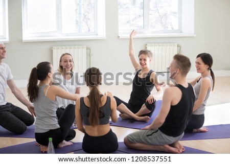 Millennial girls guys sitting together on mats in a circle laughing have fun during sport yoga seminar in professional training studio. People learn more about spiritual mental physical yoga practice Royalty-Free Stock Photo #1208537551