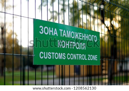 Green sign with the inscription in Russian and English: Customs control zone on the fence of metal rods