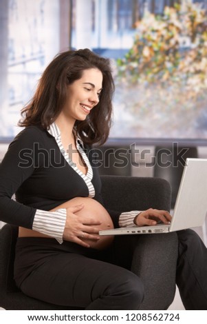 Happy young pregnant woman working in office, sitting in armchair using computer.