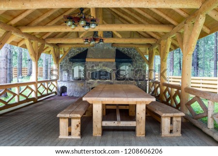 Interior of beautifully decorated modern open wooden pavilion with beam ceiling and walls, massive table, benches and big stone fireplace for barbecue and recreation. Luxurious lifestyle concept.