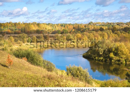 Beautiful autumn landscape with a river in a rural area . Clear blue sky with white clouds. Green and yellow trees. Purple leaves on the trees. Yellow withering grass in the fields.