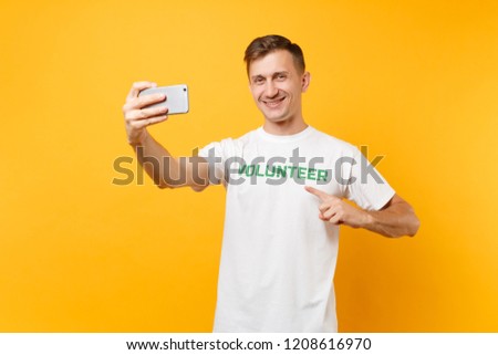 Portrait of man in white t-shirt written inscription green title volunteer taking selfie shot on mobile phone isolated on yellow background. Voluntary free assistance help charity grace work concept