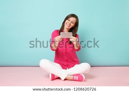 Full length woman in rose shirt, white pants sitting on floor doing selfie shot on cellphone isolated on bright pink blue pastel wall background studio. Fashion lifestyle concept. Mock up copy space