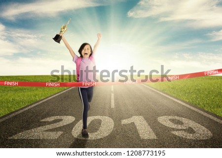 Picture of happy school girl  is carrying a trophy while crossing the success line with number 2019 on the road