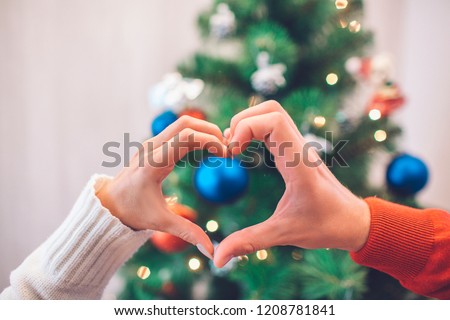 Nice picture of people's hands making hearts in front of Christmas tree with toys. There is some lights as well. Poeple wears red and white sweaters.