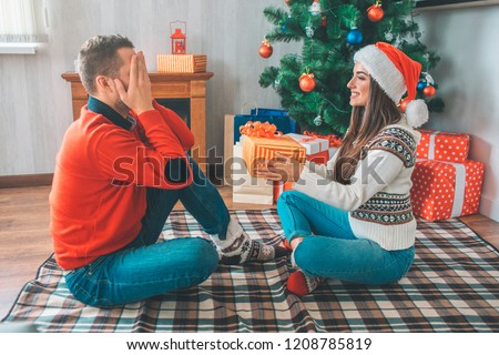 Picture of man sitting in front of woman and keeps eyes closed. She holds present for him. Young woman looks confident and cheerful.