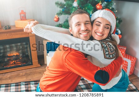 Nice and cheerful picture of young couple embrace each other. They smile. She keeps eyes closed. He looks at camera. Yoing woman keeps hands on his neck straight.