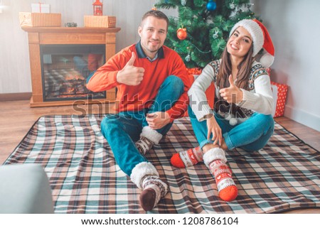 Positive picture of young couple sitting on blanket on floor and pose on camera. They keep big thumbs up and smile. People hold legs crossed.