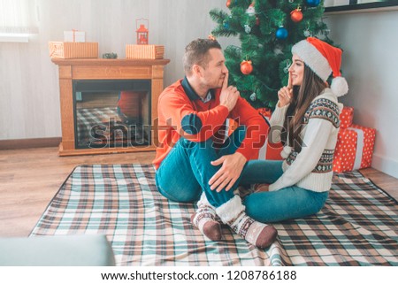 Gourgeous picture of couple. Young man and woman sitting with their legs crossed and look at each other. They keep fingers close to lips. Cheerful model smiles. Young woman laughing. Guy smiles.