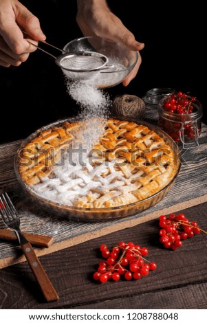 Man's hand sifting powdered sugar through sieve on round apple pie vith viburnum, wooden forks, rustic, wooden brown table