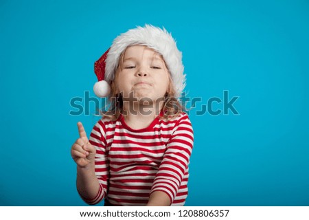 Funny little child in santa hat pointing up isolated on blue background. Funny kid playing against blue wall at home in Christmas night. Happy xmas and New Year, winter holiday concept