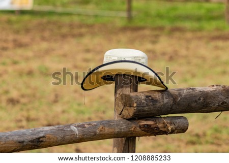 Western cowboy hat sitting on wooden fence. Italian horses and Rodeo show, surrendering sign, challenge lost. Cinematic atmosphere, suspense, dirt, grass, rural landscape and country lifestyle.  Royalty-Free Stock Photo #1208885233