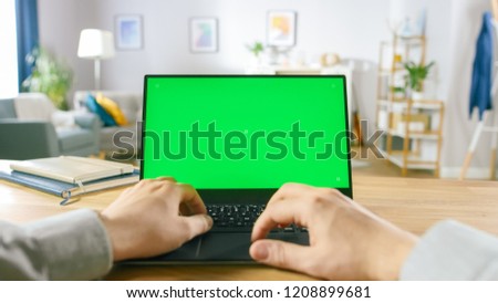 First Person Close-up Shot Man Uses Laptop with Green Mock-up Screen While Sitting at the Desk in His Cozy Living Room.