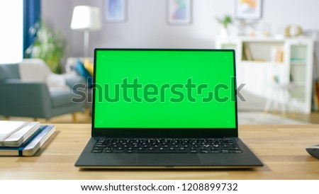 Modern Laptop with Green Mock-up Screen Display Standing on the Desk in the Cozy Living Room. Man with Mobile Phone Walks Through His Flat.