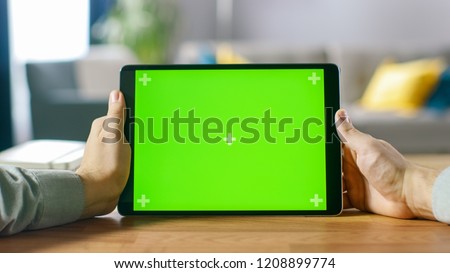 Close-up of Man Using Green Mock-up Screen Digital Tablet Computer in Landscape Mode while Sitting at His Desk. In the Background Cozy Living Room.