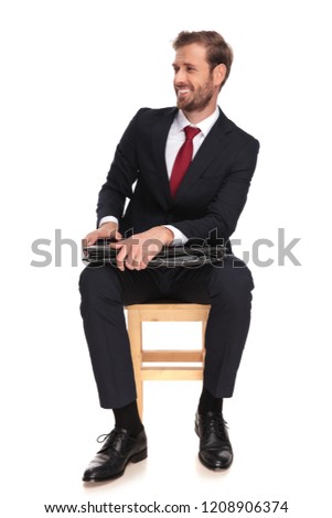 curious businessman laughing and looking to side while sitting on a wooden chair and holding his black suitcase, full length picture