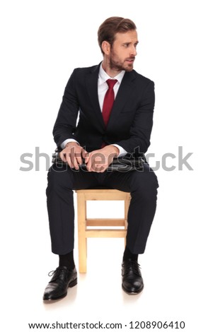 serious businessman with suitcase sits on wooden chair on white background and looks to side, full length picture