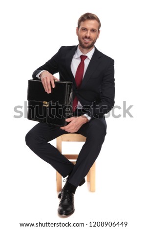 attractive businessman holding black briefcase sits on wooden chair on white background, looking relaxed, full body picture