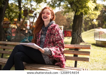 Portrait of beautiful redhead young laughing happy woman with freckles and dark lipstick wearing sunglasses reading book outdoors in park sit on bench. Looking camera.