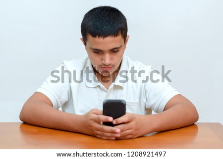 Sitting young adult brunette boy writes a message on smartphone. Teenager with phone in hand sitting at table and studying at home.