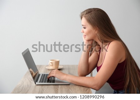 Young woman with laptop sitting at table against light background