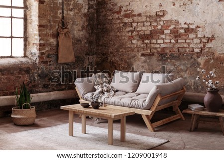 Wooden table on rug in front of beige couch in apartment interior in wabi sabi style with red brick wall. Real photo