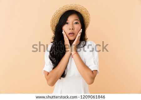 Portrait of a shocked asian girl in summer hat isolated over beige background, holding hands at her face