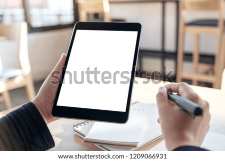 Mockup image of hands holding and using black tablet pc with blank white desktop screen while writing on notebooks in office