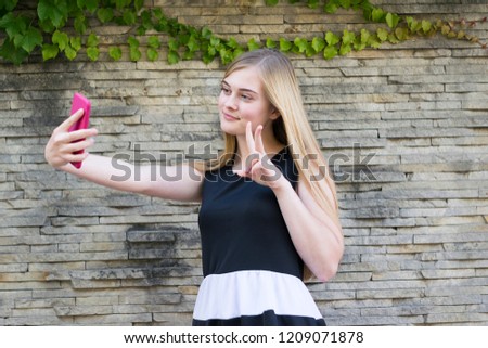 Waist-up portrait of blonde good-looking young female posing outside and taking selfie on trendy mobile phone on brick wall. Technology and lifestyle concept