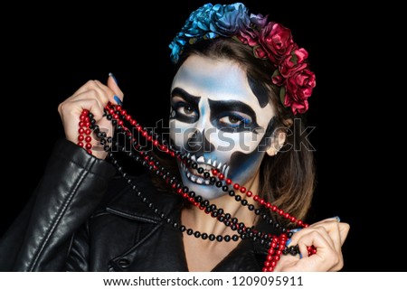 Creative makeup for Halloween party. Voodoo girl with picture makeup on her face, spooky scary skull. Holds beads jewelry. White teeth, black eyebrows drawn. Wreath of flowers, cool leather costume